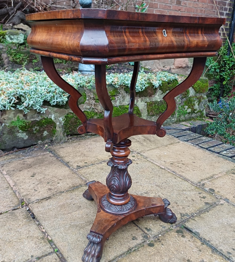A Victorian flame mahogany work table featuring a shaped top with a central keyhole escutcheon, supported by C-scroll arms, a carved acanthus pedestal, and a triform base with claw feet. View II