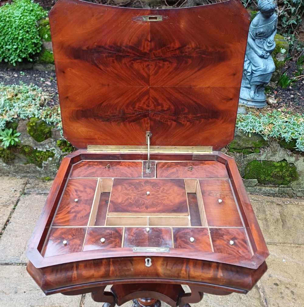 A Victorian flame mahogany work table featuring a shaped top with a central keyhole escutcheon, supported by C-scroll arms, a carved acanthus pedestal, and a triform base with claw feet. View V