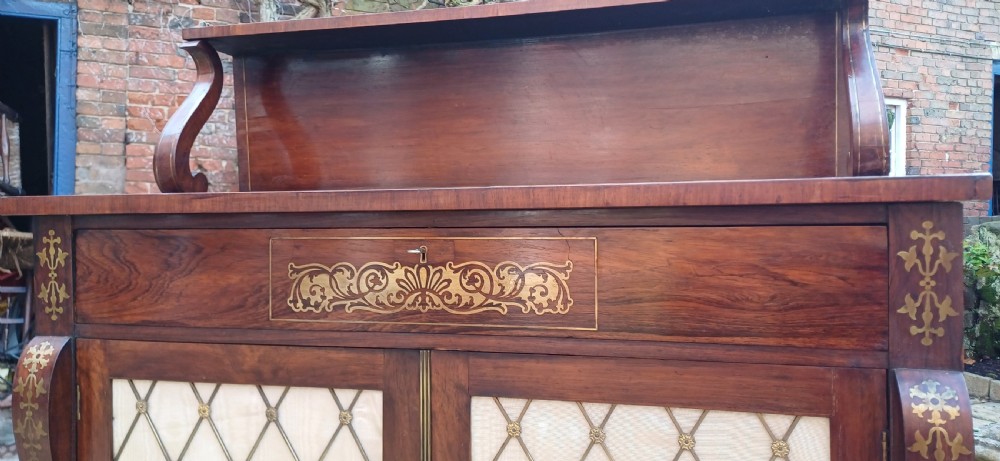 A Regency rosewood chiffonier featuring decorative brass inlay, a raised back shelf with a brass gallery, and two cupboard doors with brass wire lattice grilles over silk. View VIII