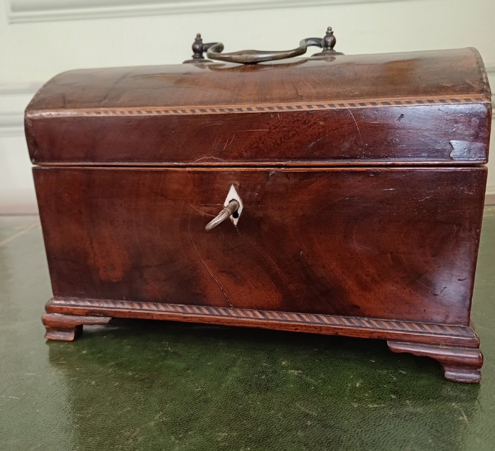 An antique Georgian mahogany tea caddy featuring a domed lid, brass top handle, chevron string inlay, and a diamond bone escutcheon. View VI