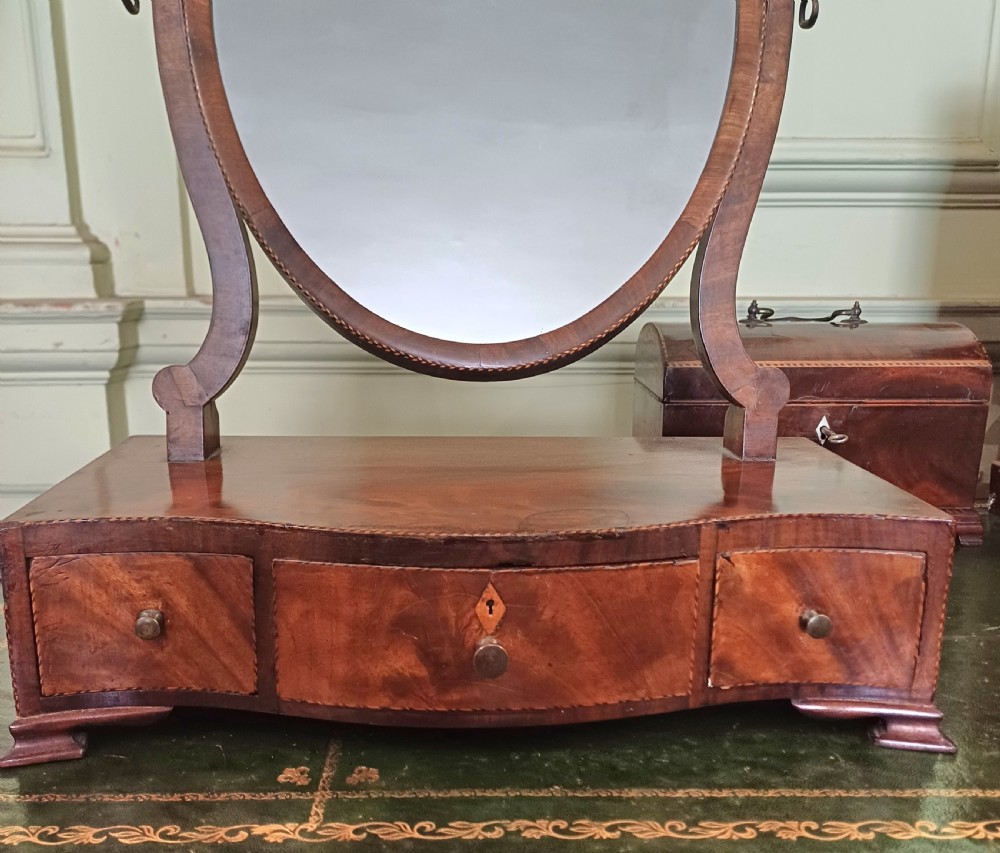 A Georgian flame mahogany serpentine toilet mirror featuring an oval adjustable mirror on curved supports set above a three-drawer box base. View II