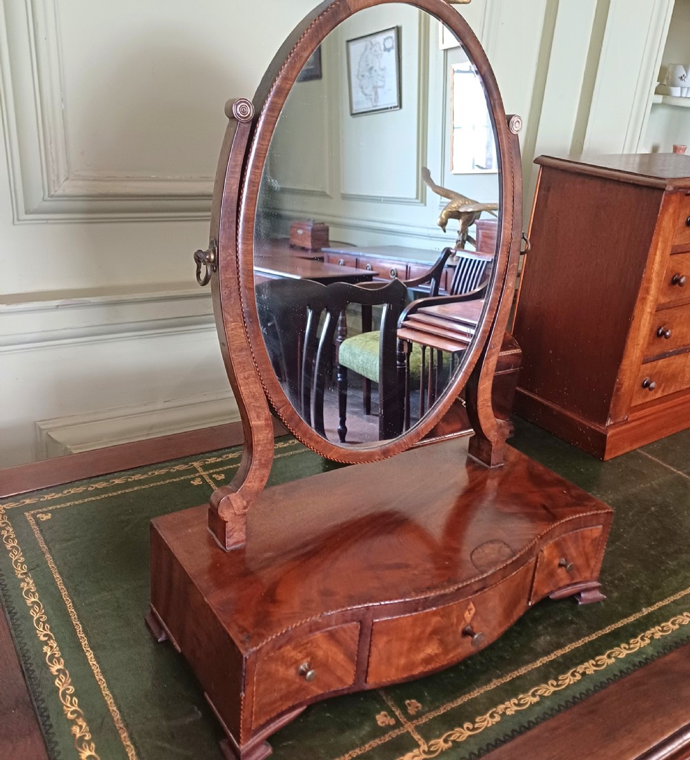 A Georgian flame mahogany serpentine toilet mirror featuring an oval adjustable mirror on curved supports set above a three-drawer box base. View IV