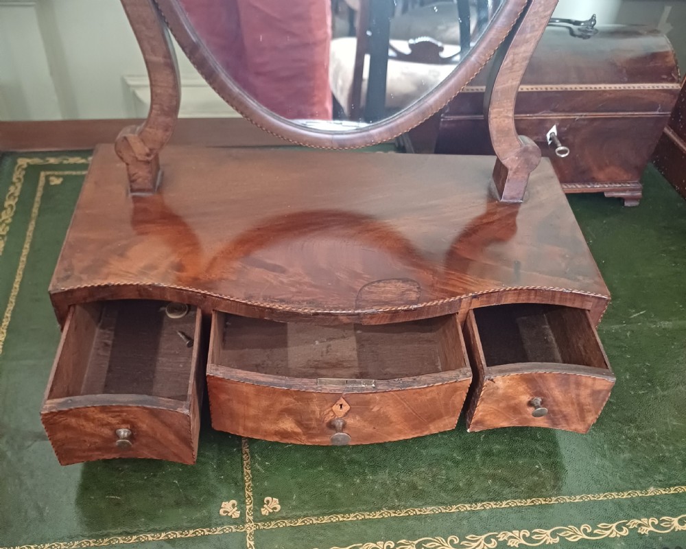 A Georgian flame mahogany serpentine toilet mirror featuring an oval adjustable mirror on curved supports set above a three-drawer box base. View VII