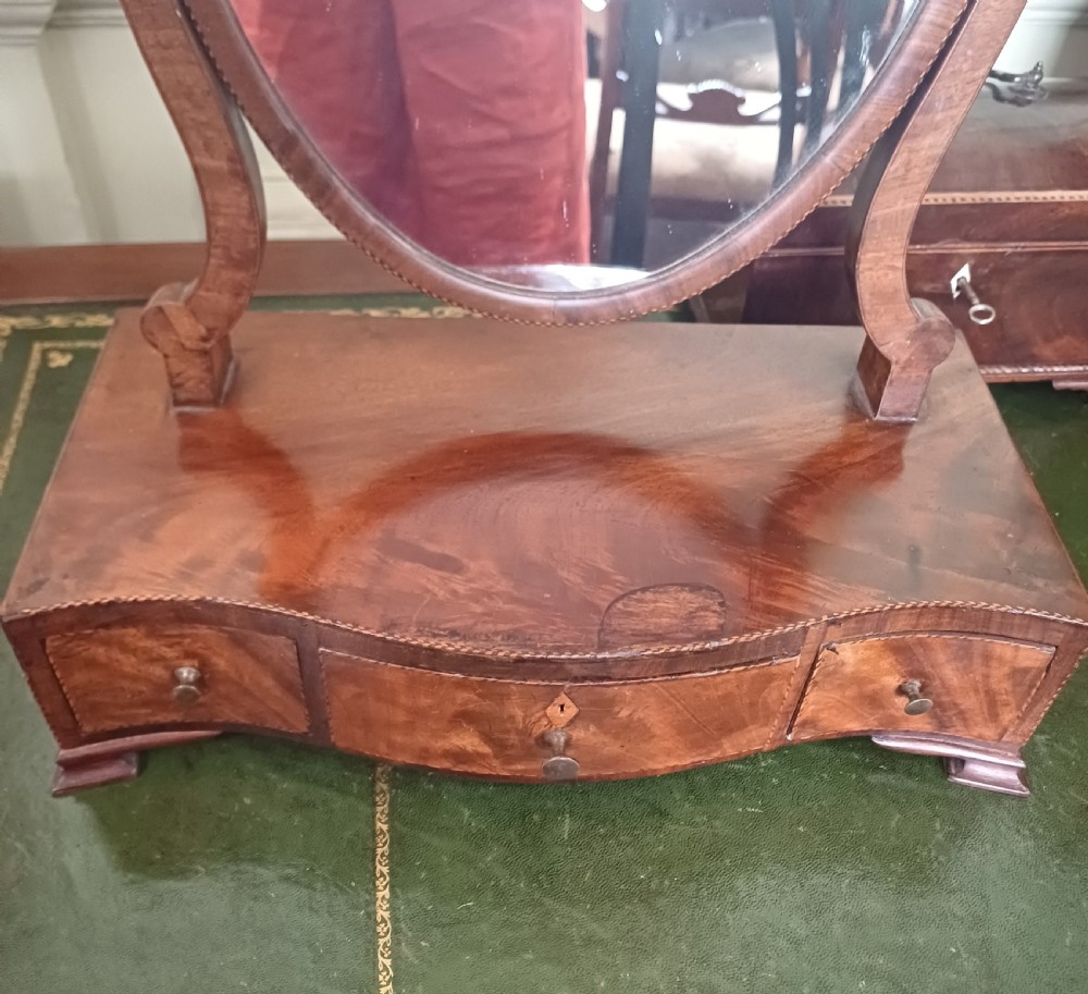 A Georgian flame mahogany serpentine toilet mirror featuring an oval adjustable mirror on curved supports set above a three-drawer box base. View VIII
