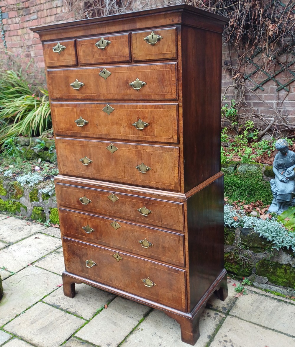 An early 18th-century walnut chest on chest featuring multiple tiered drawers, crossbanded inlay, brass drop handles, and bracket feet. View II