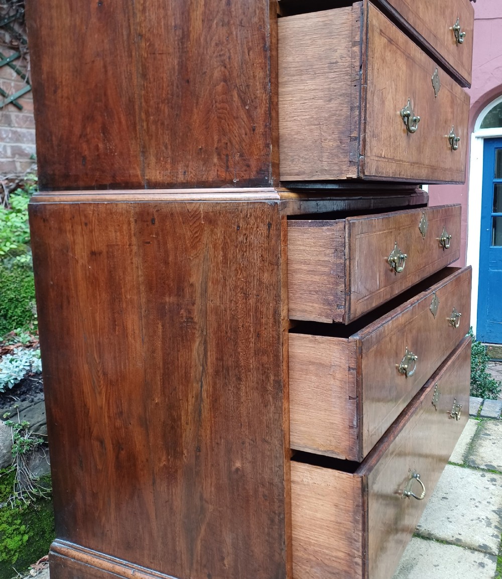 An early 18th-century walnut chest on chest featuring multiple tiered drawers, crossbanded inlay, brass drop handles, and bracket feet. View V