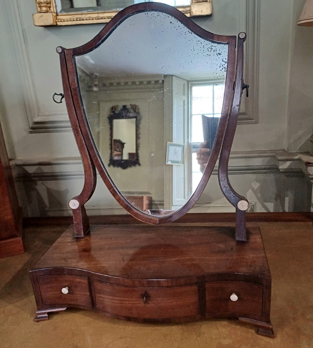 A Georgian mahogany toilet mirror featuring a serpentine-fronted base with three drawers and a central pivoting mirror supported by curved uprights. View II