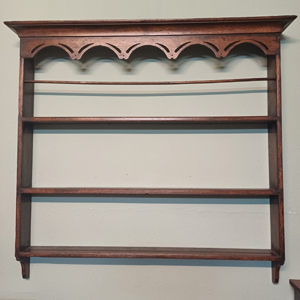 An 18th-century dark oak wall-mounted plate rack featuring three horizontal open display shelves, a protective retainage rail, and a shaped arch frieze at the top.