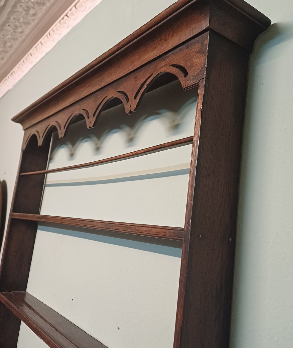 An 18th-century dark oak wall-mounted plate rack featuring three horizontal open display shelves, a protective retainage rail, and a shaped arch frieze at the top. View II