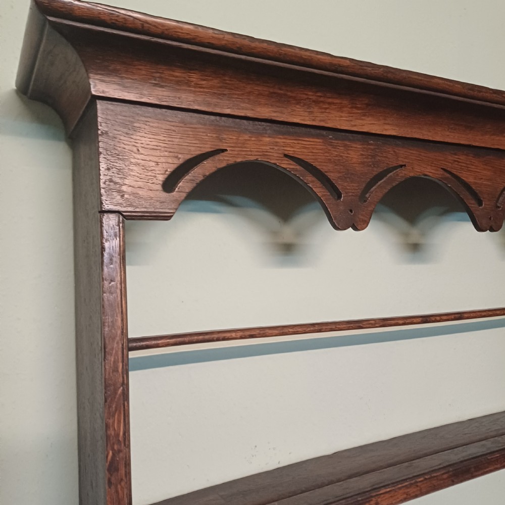 An 18th-century dark oak wall-mounted plate rack featuring three horizontal open display shelves, a protective retainage rail, and a shaped arch frieze at the top. View IV