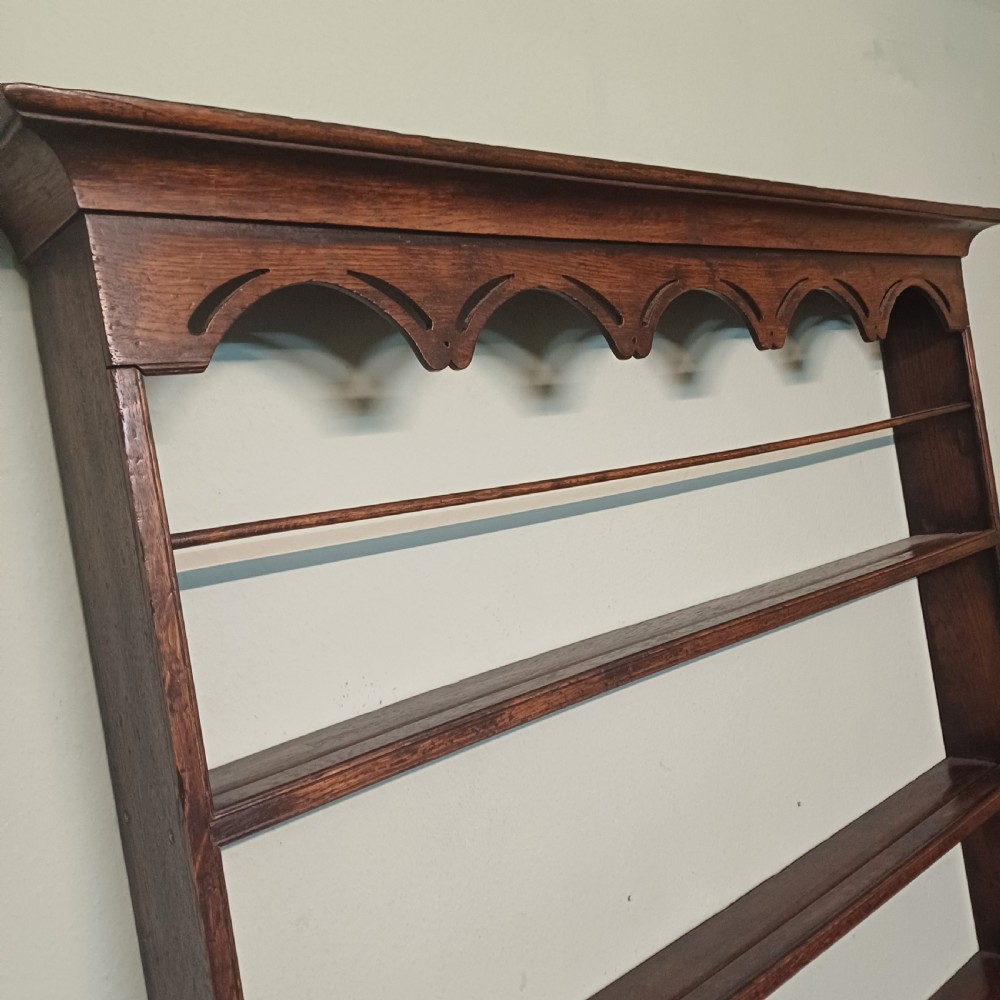 An 18th-century dark oak wall-mounted plate rack featuring three horizontal open display shelves, a protective retainage rail, and a shaped arch frieze at the top. View V