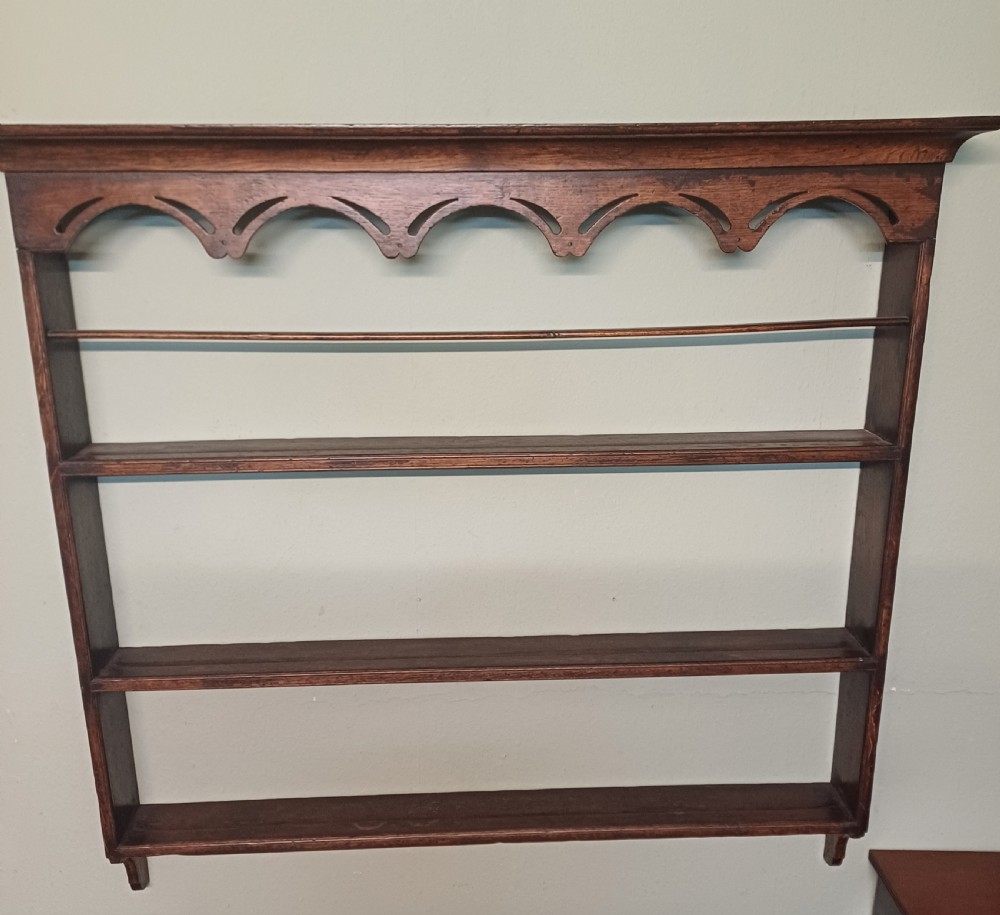 An 18th-century dark oak wall-mounted plate rack featuring three horizontal open display shelves, a protective retainage rail, and a shaped arch frieze at the top. View VI