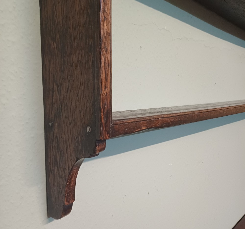 An 18th-century dark oak wall-mounted plate rack featuring three horizontal open display shelves, a protective retainage rail, and a shaped arch frieze at the top. View VII