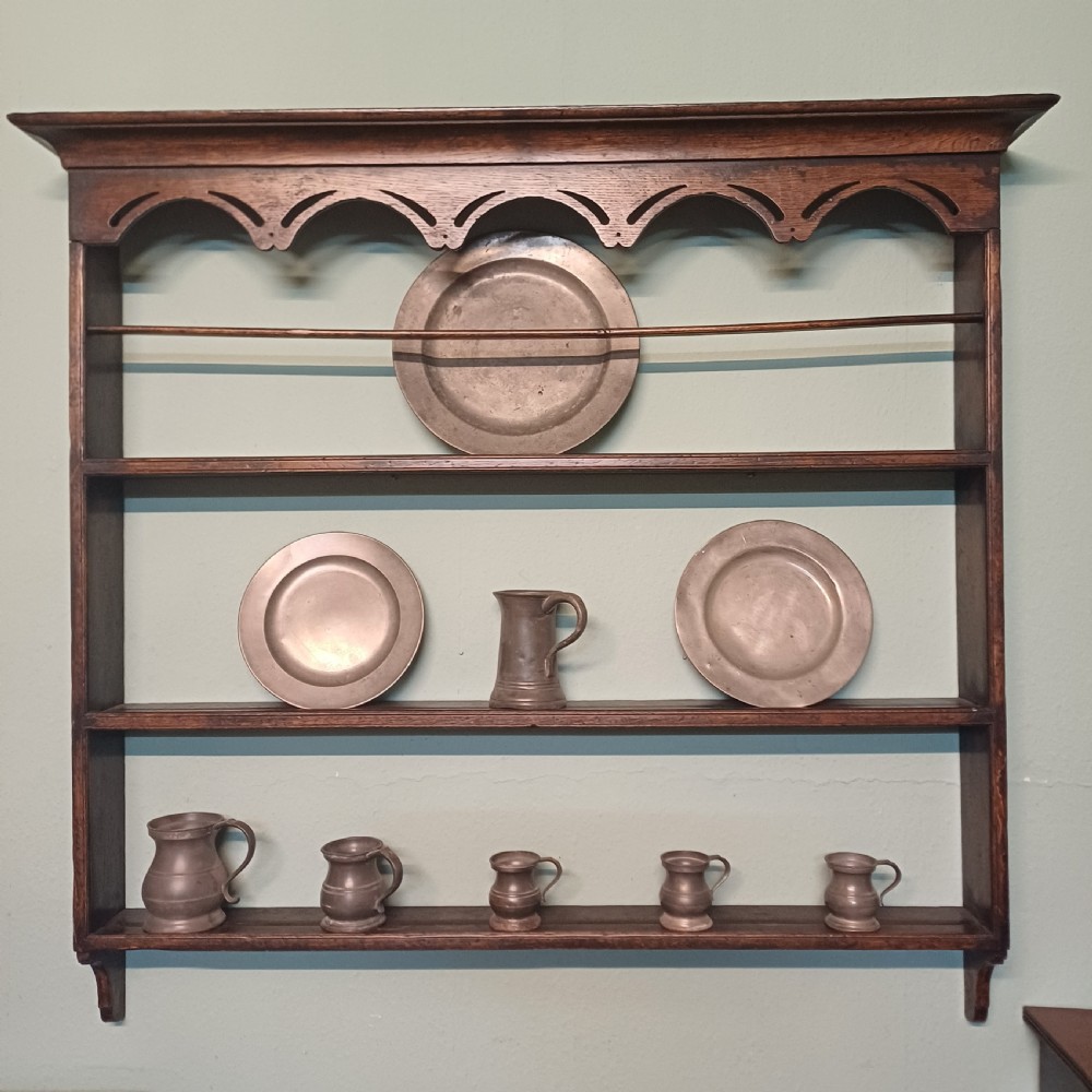 An 18th-century dark oak wall-mounted plate rack featuring three horizontal open display shelves, a protective retainage rail, and a shaped arch frieze at the top. View VIII