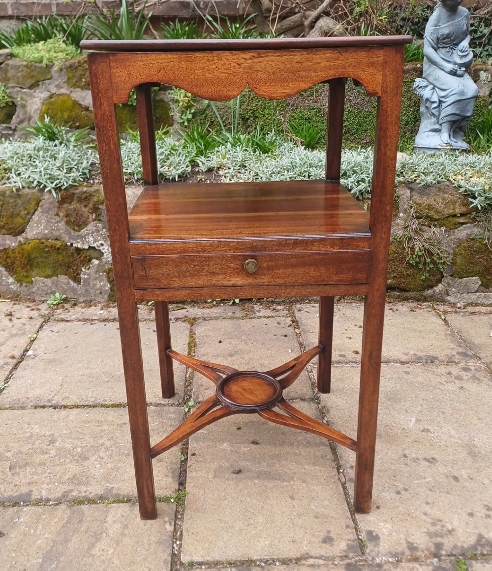 An antique Georgian mahogany bedside table featuring an open top tier with a scalloped apron, a single drawer with a brass pull, square legs, and an X-shaped stretcher with a central circular platform.