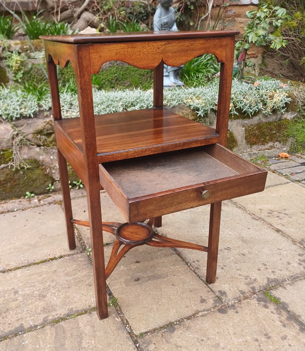 An antique Georgian mahogany bedside table featuring an open top tier with a scalloped apron, a single drawer with a brass pull, square legs, and an X-shaped stretcher with a central circular platform. View II
