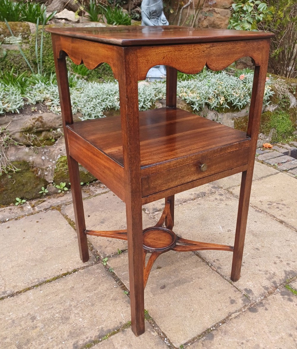 An antique Georgian mahogany bedside table featuring an open top tier with a scalloped apron, a single drawer with a brass pull, square legs, and an X-shaped stretcher with a central circular platform. View III
