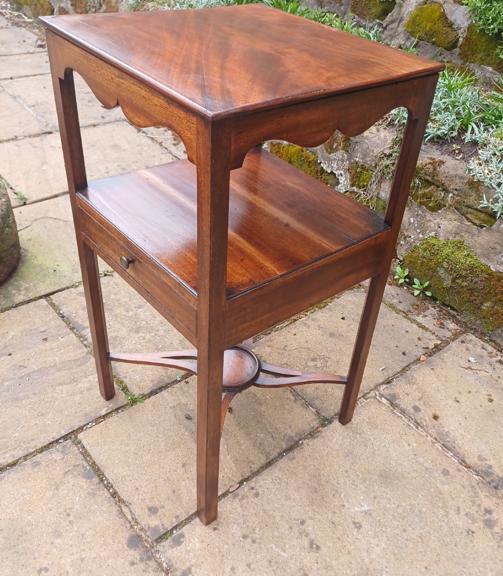 An antique Georgian mahogany bedside table featuring an open top tier with a scalloped apron, a single drawer with a brass pull, square legs, and an X-shaped stretcher with a central circular platform. View IV