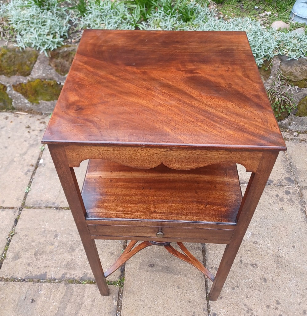 An antique Georgian mahogany bedside table featuring an open top tier with a scalloped apron, a single drawer with a brass pull, square legs, and an X-shaped stretcher with a central circular platform. View V