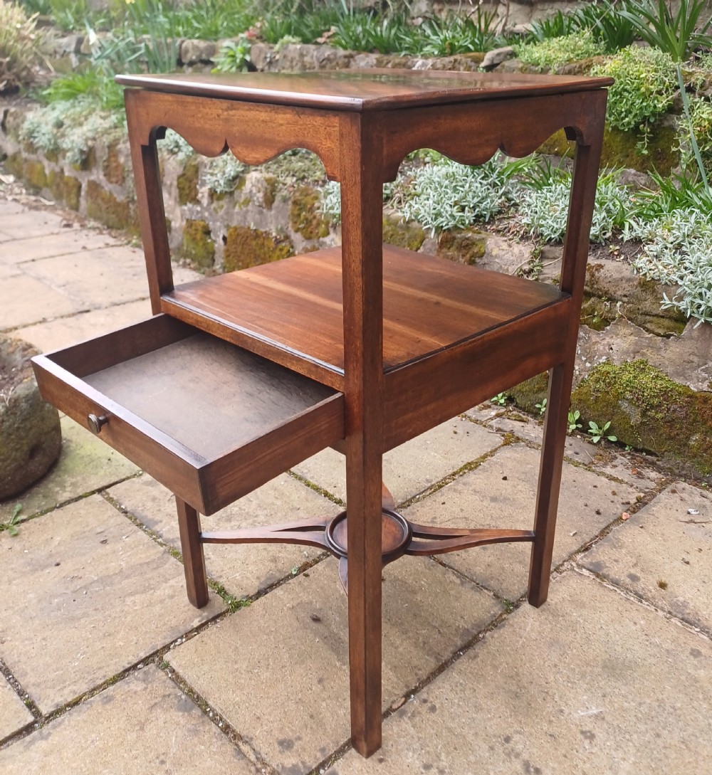 An antique Georgian mahogany bedside table featuring an open top tier with a scalloped apron, a single drawer with a brass pull, square legs, and an X-shaped stretcher with a central circular platform. View VI