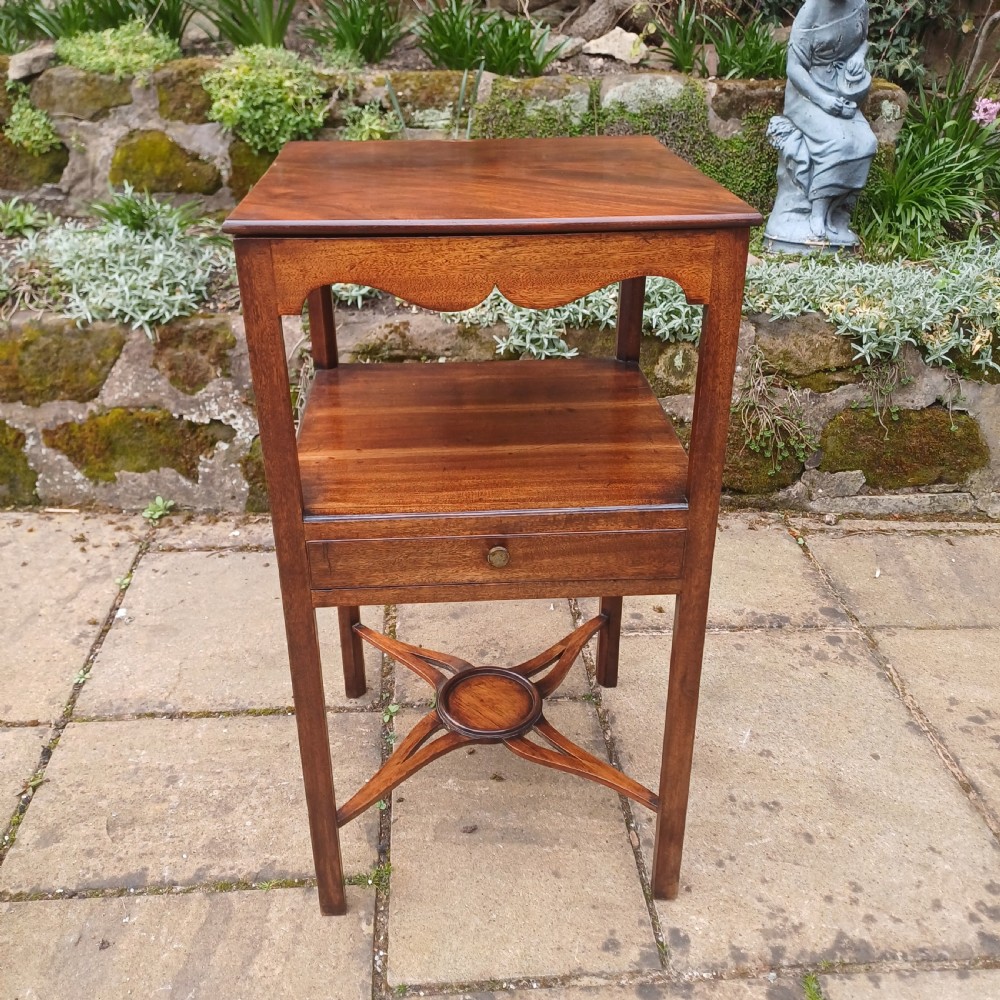 An antique Georgian mahogany bedside table featuring an open top tier with a scalloped apron, a single drawer with a brass pull, square legs, and an X-shaped stretcher with a central circular platform. View IX