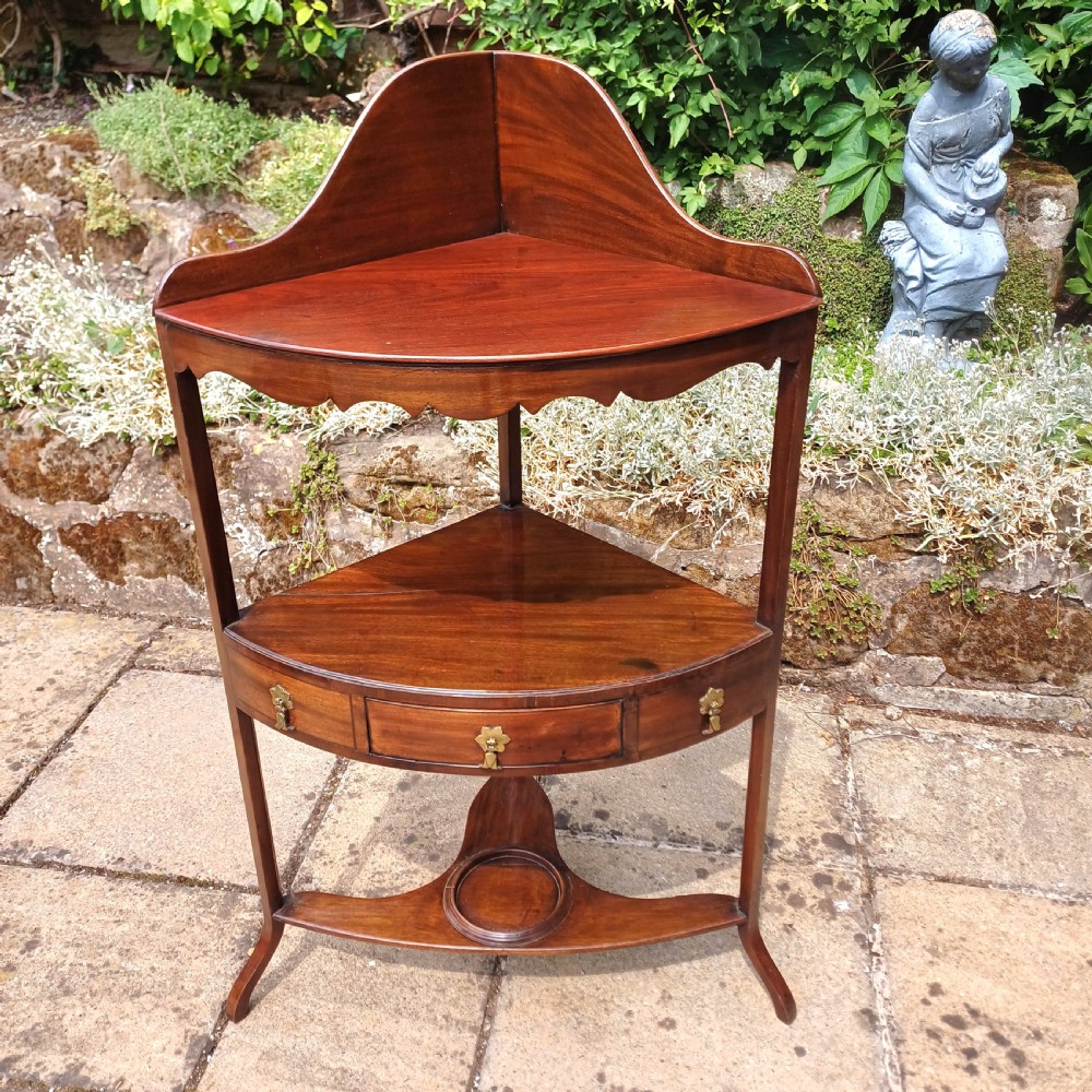 A Georgian mahogany corner washstand featuring a curved splashback, middle drawer with brass handles, and lower stretcher with a basin ring.