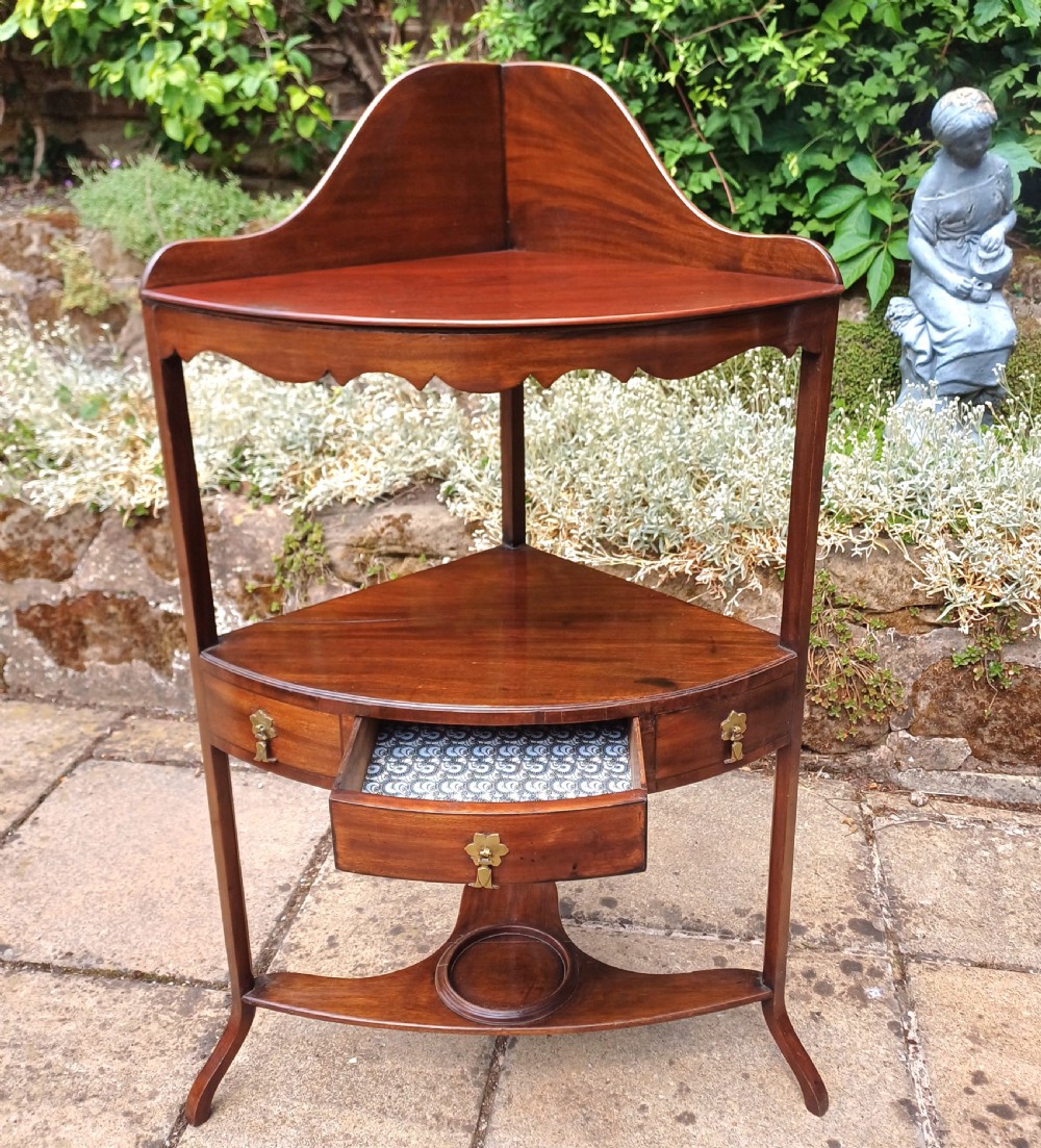 A Georgian mahogany corner washstand featuring a curved splashback, middle drawer with brass handles, and lower stretcher with a basin ring. View VI