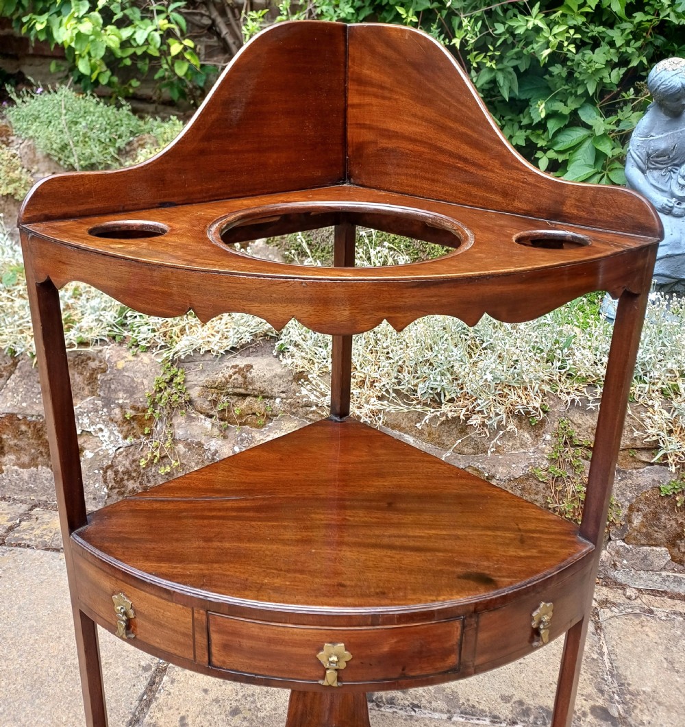 A Georgian mahogany corner washstand featuring a curved splashback, middle drawer with brass handles, and lower stretcher with a basin ring. View XI