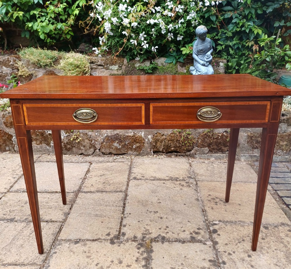 An Edwardian mahogany writing table featuring two inlaid drawers, brass oval drop handles, and slender tapered legs.