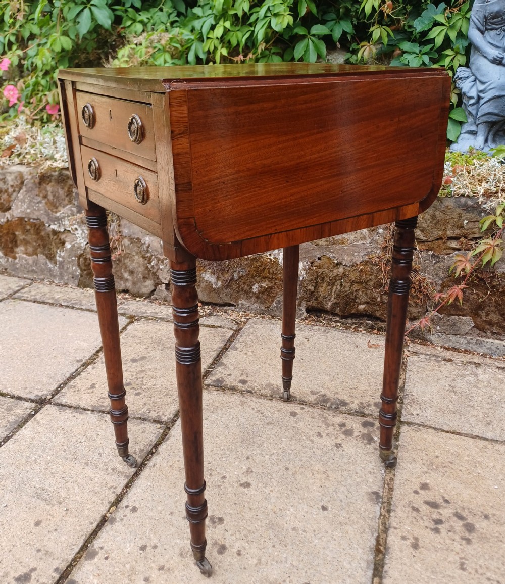A Regency mahogany miniature Pembroke table featuring two drop leaves, two drawers with brass ring handles, and turned reeded legs on castors. View X
