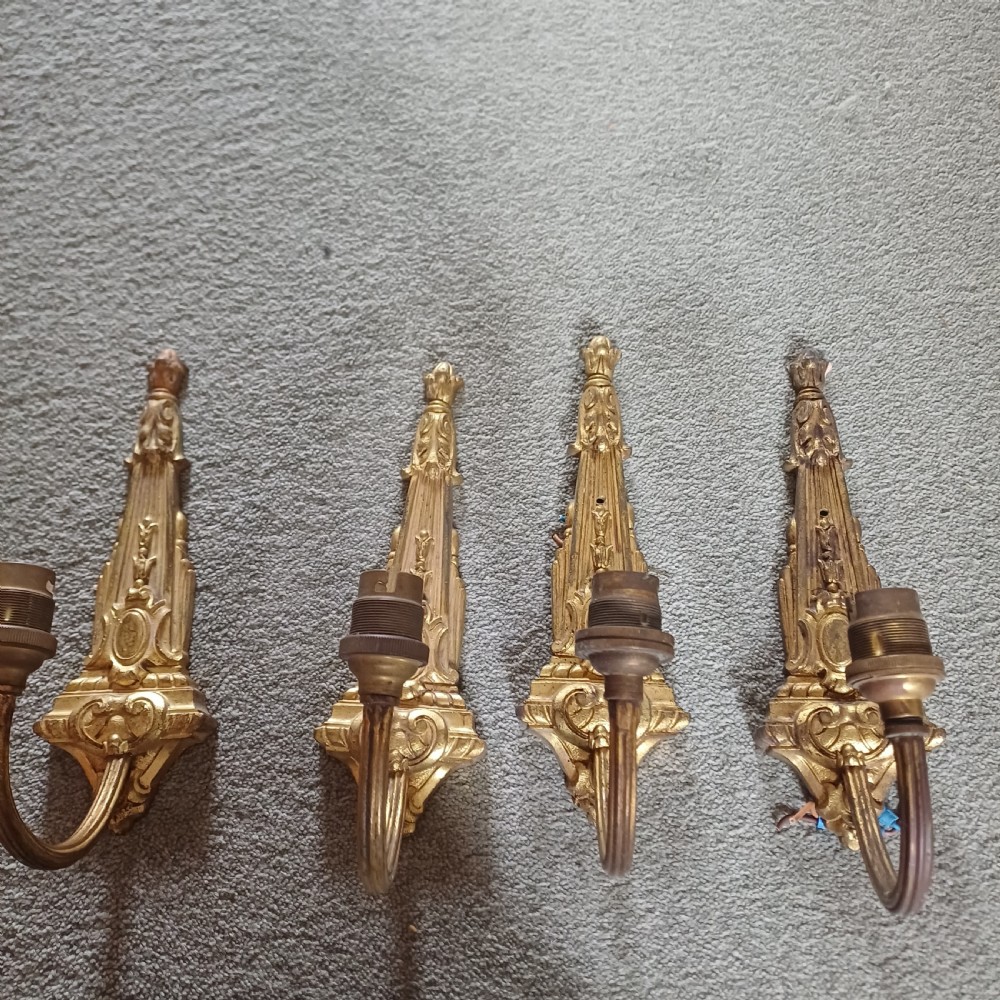 A set of four ornate gilded brass single-arm wall sconces featuring tapered backplates with classical scroll detailing.