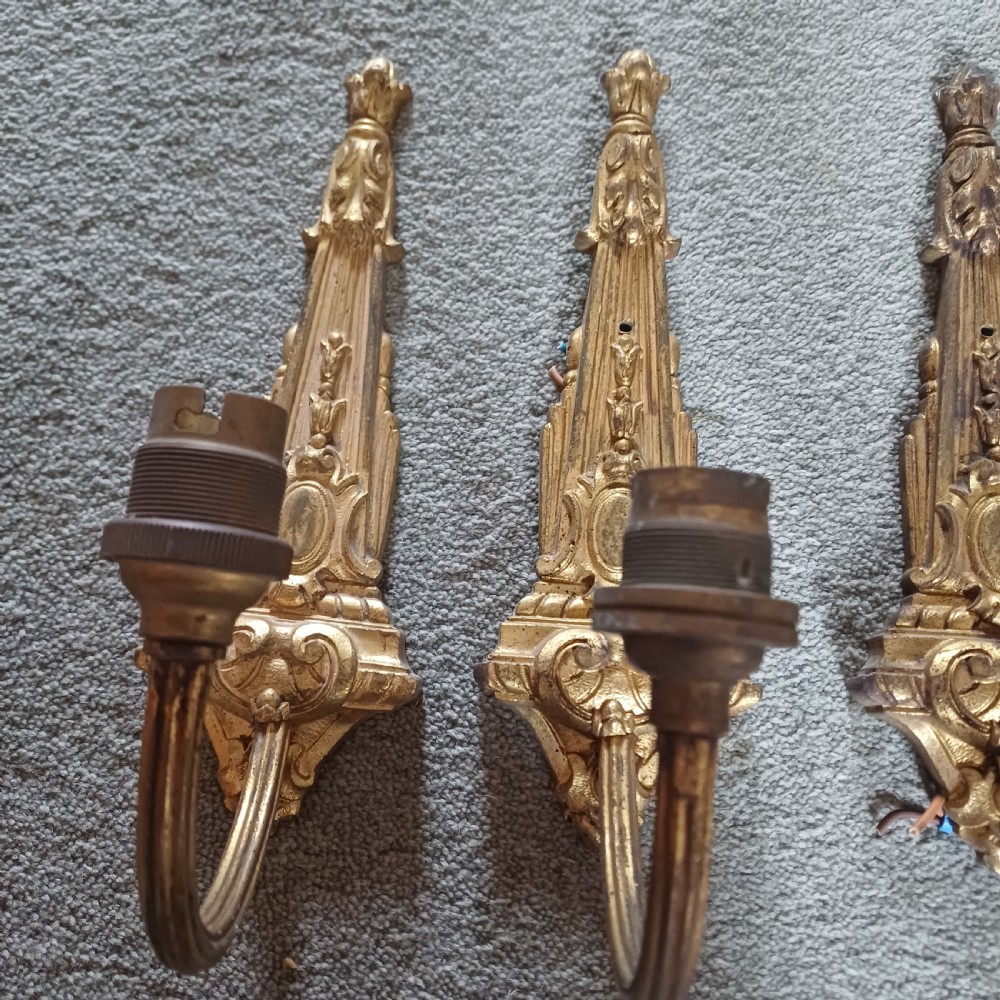 A set of four ornate gilded brass single-arm wall sconces featuring tapered backplates with classical scroll detailing. View II