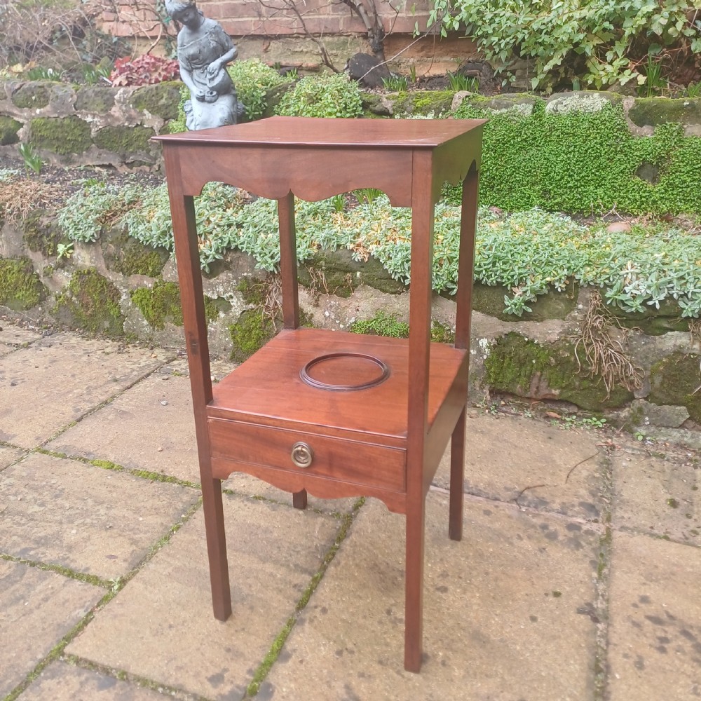 A two-tier Georgian mahogany bedside table featuring a square top, a lower shelf with a circular inset recess, a single drawer with a brass ring handle, and scalloped aprons.