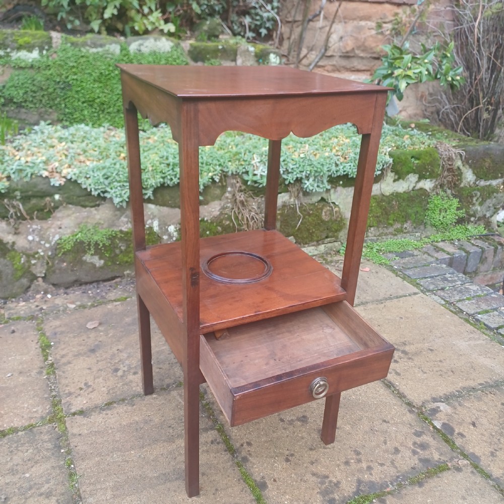 A two-tier Georgian mahogany bedside table featuring a square top, a lower shelf with a circular inset recess, a single drawer with a brass ring handle, and scalloped aprons. View II