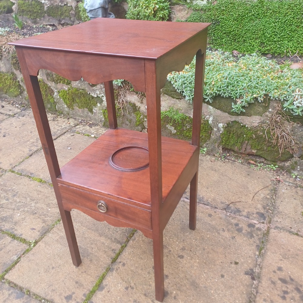 A two-tier Georgian mahogany bedside table featuring a square top, a lower shelf with a circular inset recess, a single drawer with a brass ring handle, and scalloped aprons. View III
