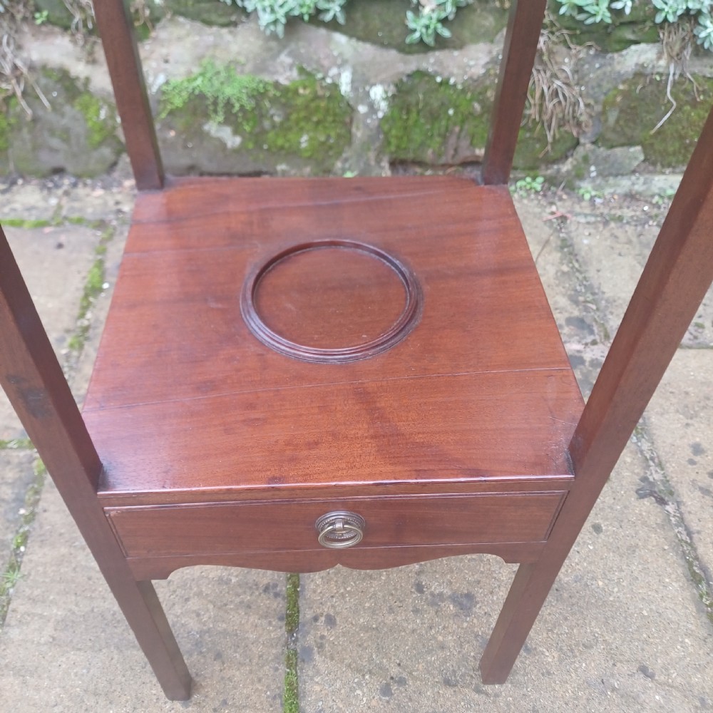 A two-tier Georgian mahogany bedside table featuring a square top, a lower shelf with a circular inset recess, a single drawer with a brass ring handle, and scalloped aprons. View IV