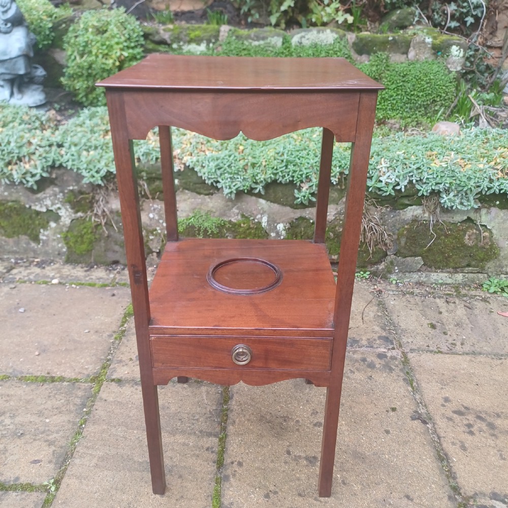 A two-tier Georgian mahogany bedside table featuring a square top, a lower shelf with a circular inset recess, a single drawer with a brass ring handle, and scalloped aprons. View VI