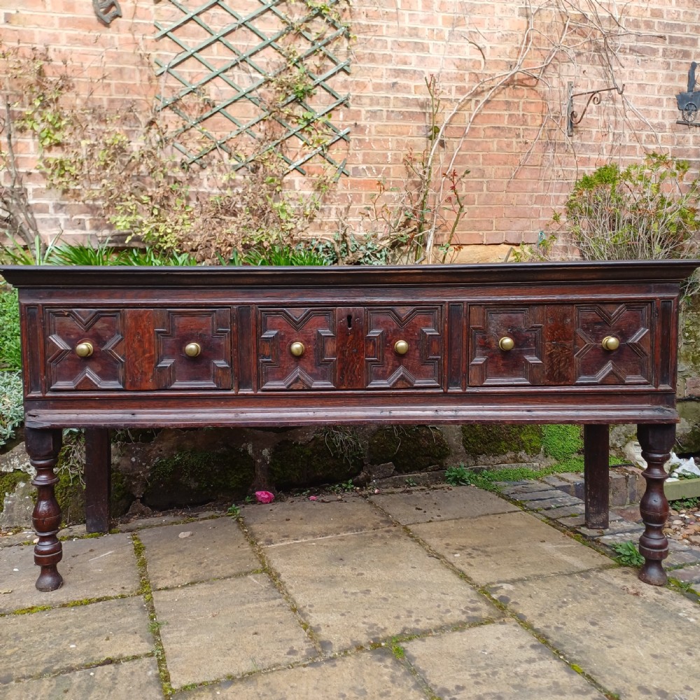 A late seventeenth-century dark oak dresser base featuring geometric mitred moulding across three drawers, turned front legs, and brass knob handles.