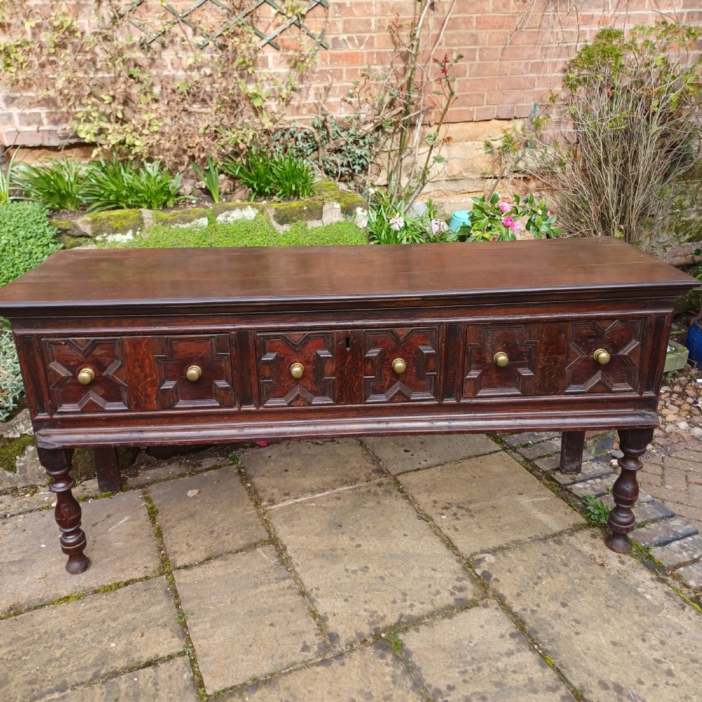 A late seventeenth-century dark oak dresser base featuring geometric mitred moulding across three drawers, turned front legs, and brass knob handles. View II