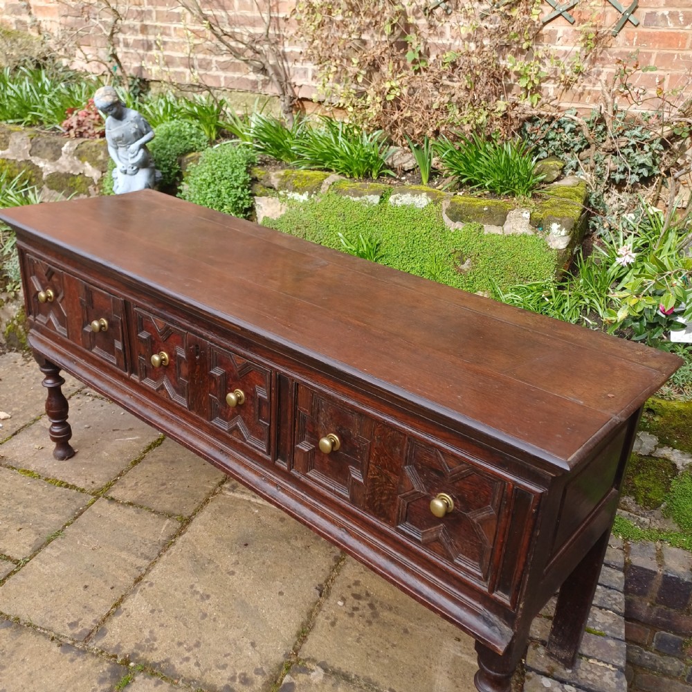 A late seventeenth-century dark oak dresser base featuring geometric mitred moulding across three drawers, turned front legs, and brass knob handles. View III