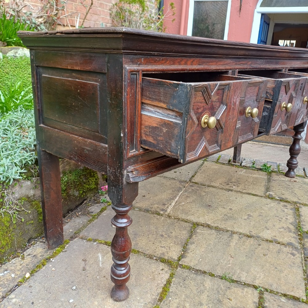 A late seventeenth-century dark oak dresser base featuring geometric mitred moulding across three drawers, turned front legs, and brass knob handles. View IV