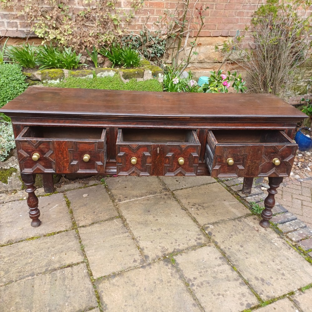 A late seventeenth-century dark oak dresser base featuring geometric mitred moulding across three drawers, turned front legs, and brass knob handles. View V