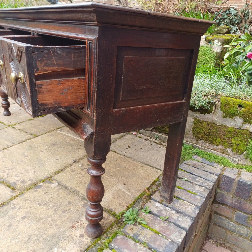 A late seventeenth-century dark oak dresser base featuring geometric mitred moulding across three drawers, turned front legs, and brass knob handles. View VI