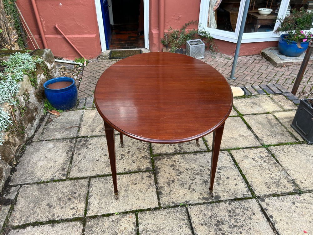 An 18th-century oval mahogany Pembroke table featuring two drop leaves, a single drawer with brass ring handles, line inlay, and square tapered legs on castors. View IV