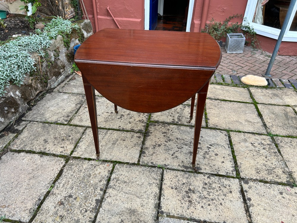 An 18th-century oval mahogany Pembroke table featuring two drop leaves, a single drawer with brass ring handles, line inlay, and square tapered legs on castors. View V
