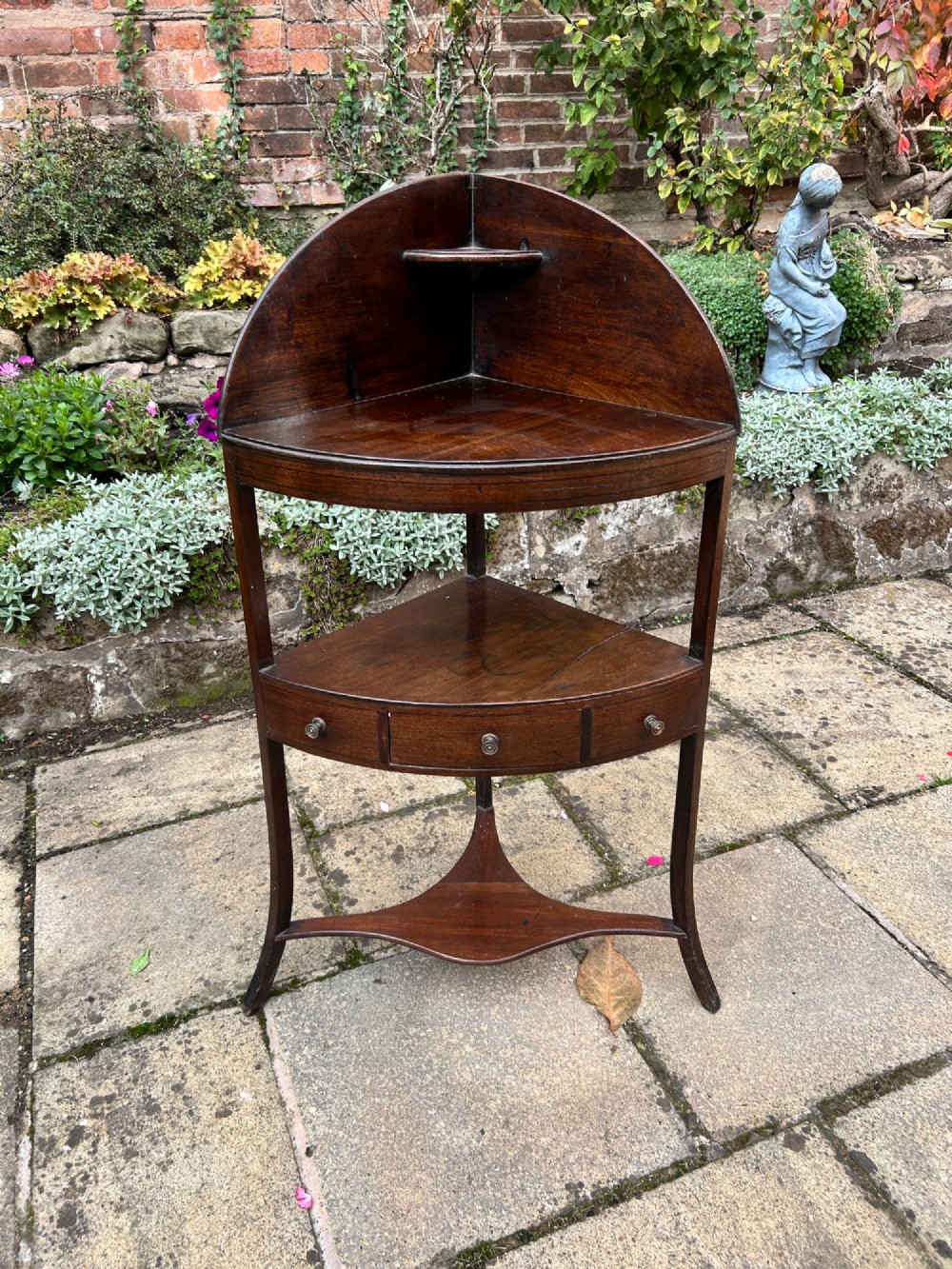 A late 18th-century mahogany corner washstand featuring a curved splashback with a small upper shelf, a middle drawer tier, and splayed legs connected by a shaped lower shelf.