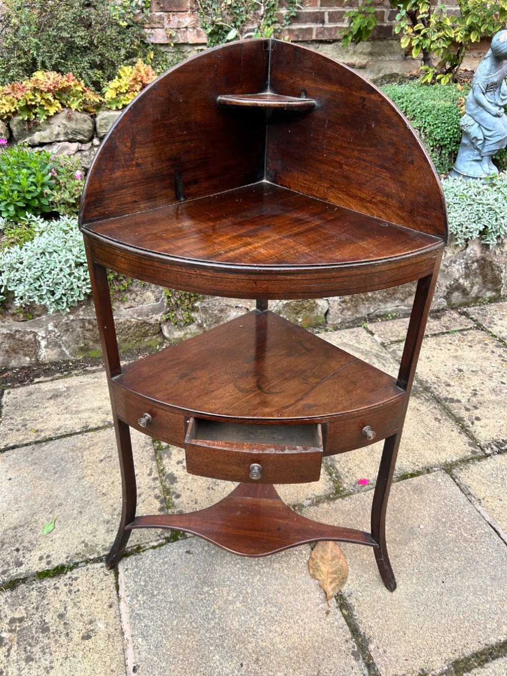 A late 18th-century mahogany corner washstand featuring a curved splashback with a small upper shelf, a middle drawer tier, and splayed legs connected by a shaped lower shelf. View II