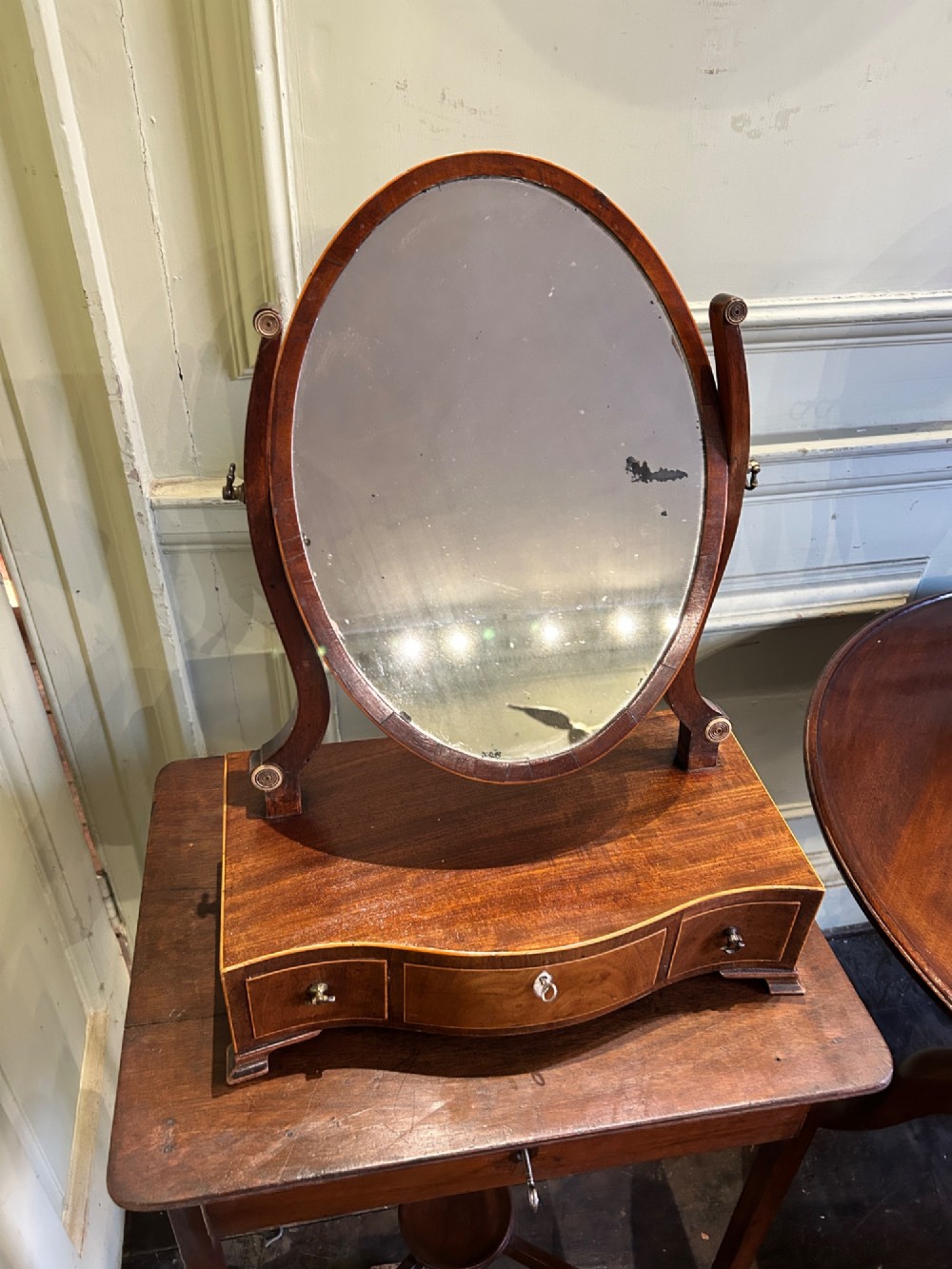 A Georgian mahogany toilet mirror featuring an oval adjustable mirror supported by curved uprights, set upon a serpentine base with three small inlaid drawers and bracket feet.