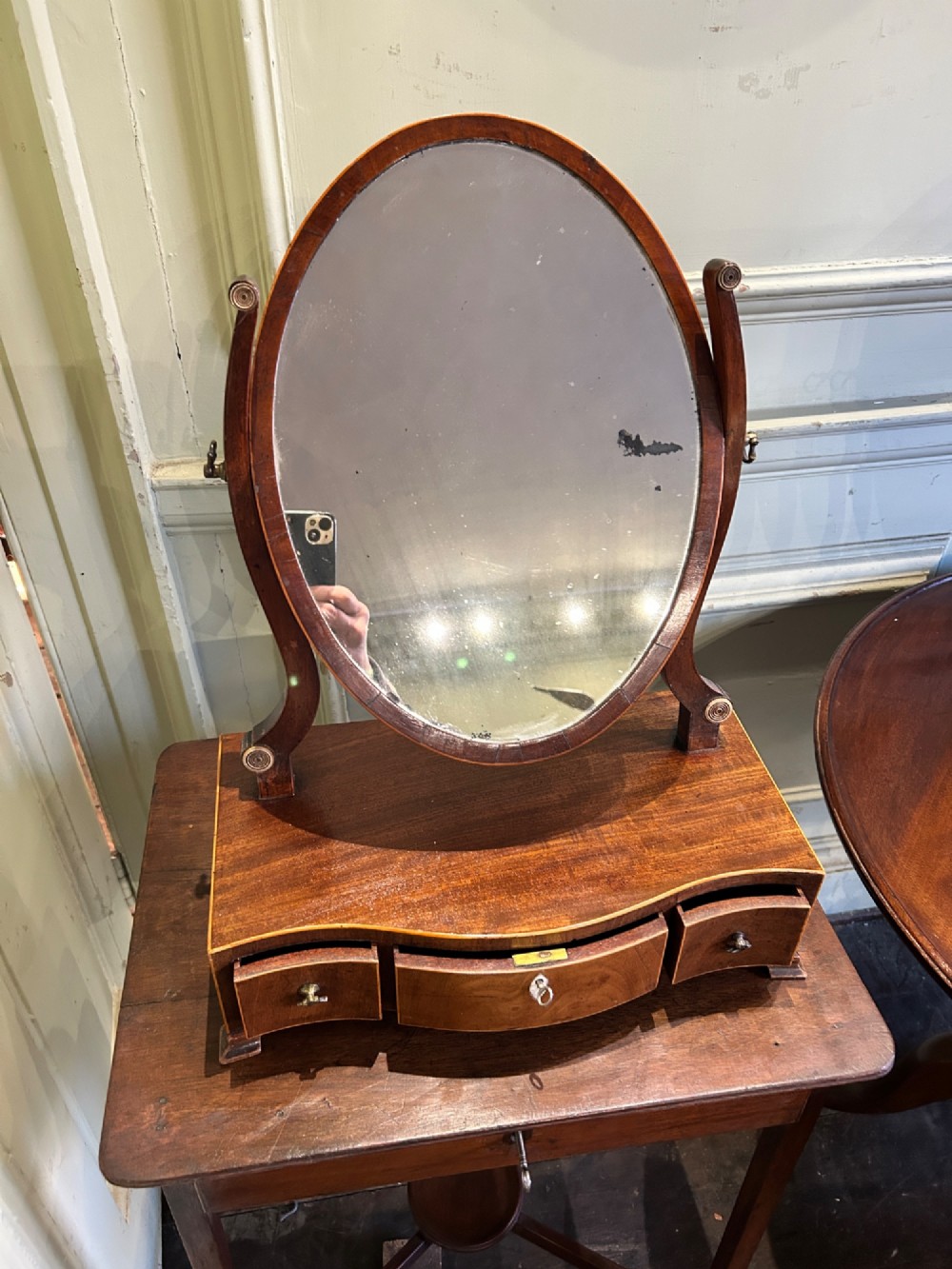 A Georgian mahogany toilet mirror featuring an oval adjustable mirror supported by curved uprights, set upon a serpentine base with three small inlaid drawers and bracket feet. View II