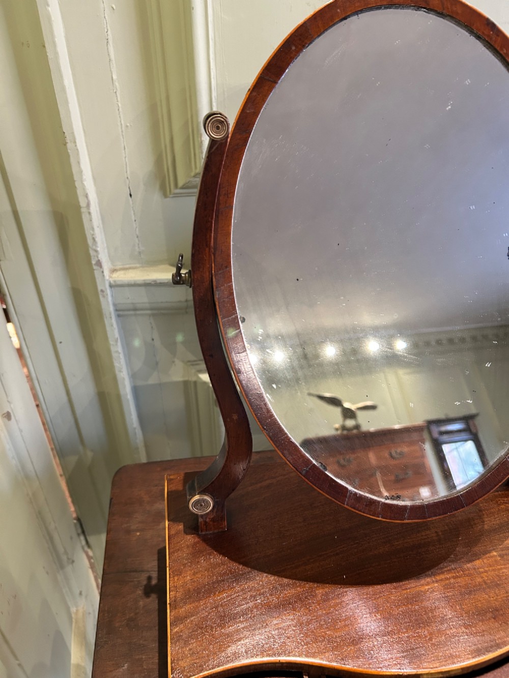 A Georgian mahogany toilet mirror featuring an oval adjustable mirror supported by curved uprights, set upon a serpentine base with three small inlaid drawers and bracket feet. View III
